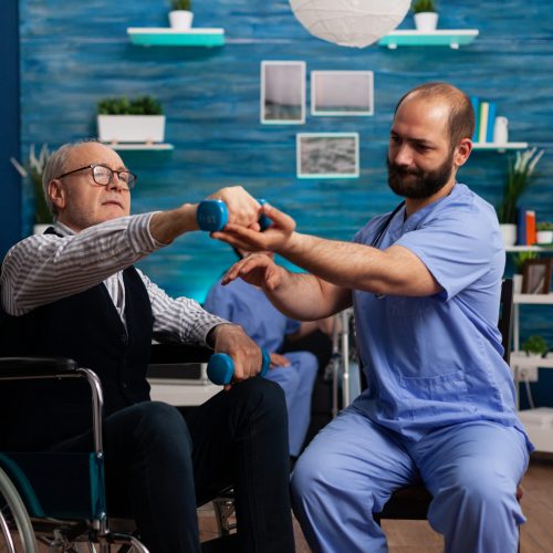 Practitioner man doctor helping retiree senior male in wheelchair to do physiotherapy strength exercise using dumbbell. Social nursing services at home. Healthcare recovery. Support worker helper